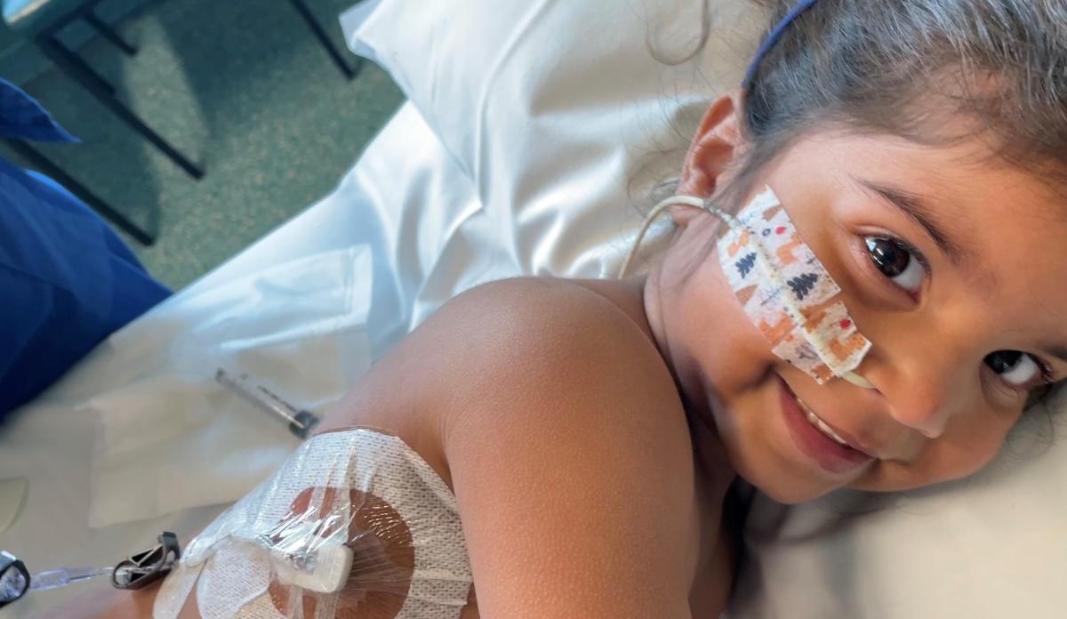Six year old Zara, in hospital bed, smiling at the camera, a feeding tube in her nose and medical ports in her side.